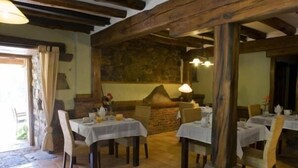 A rustic dining area with stone walls, wooden beams, and tiled flooring. Tables are set with white tablecloths and chairs.