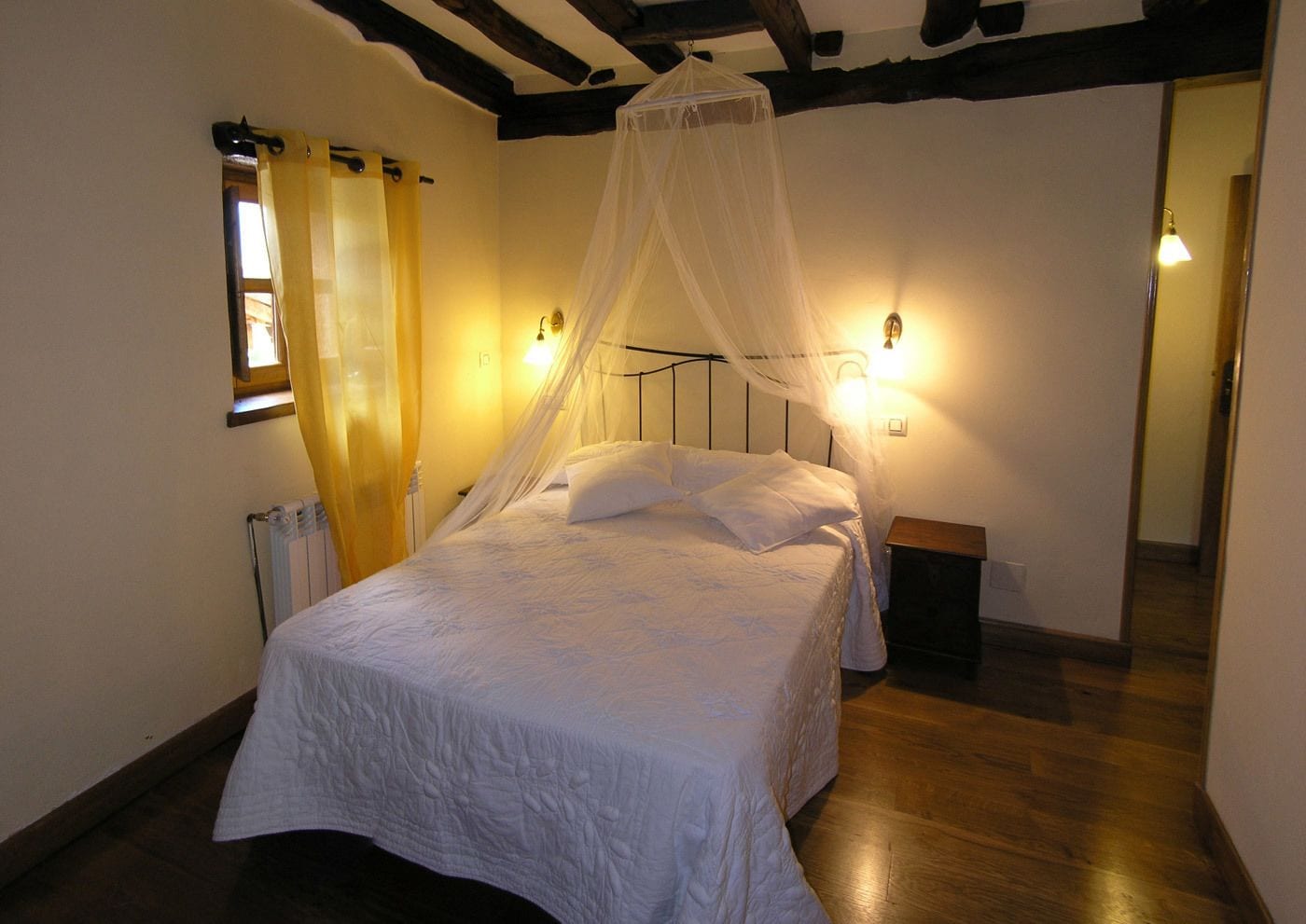 Une chambre à coucher avec un lit sous un baldaquin, une table de chevet, une commode, un radiateur et une fenêtre avec des rideaux.