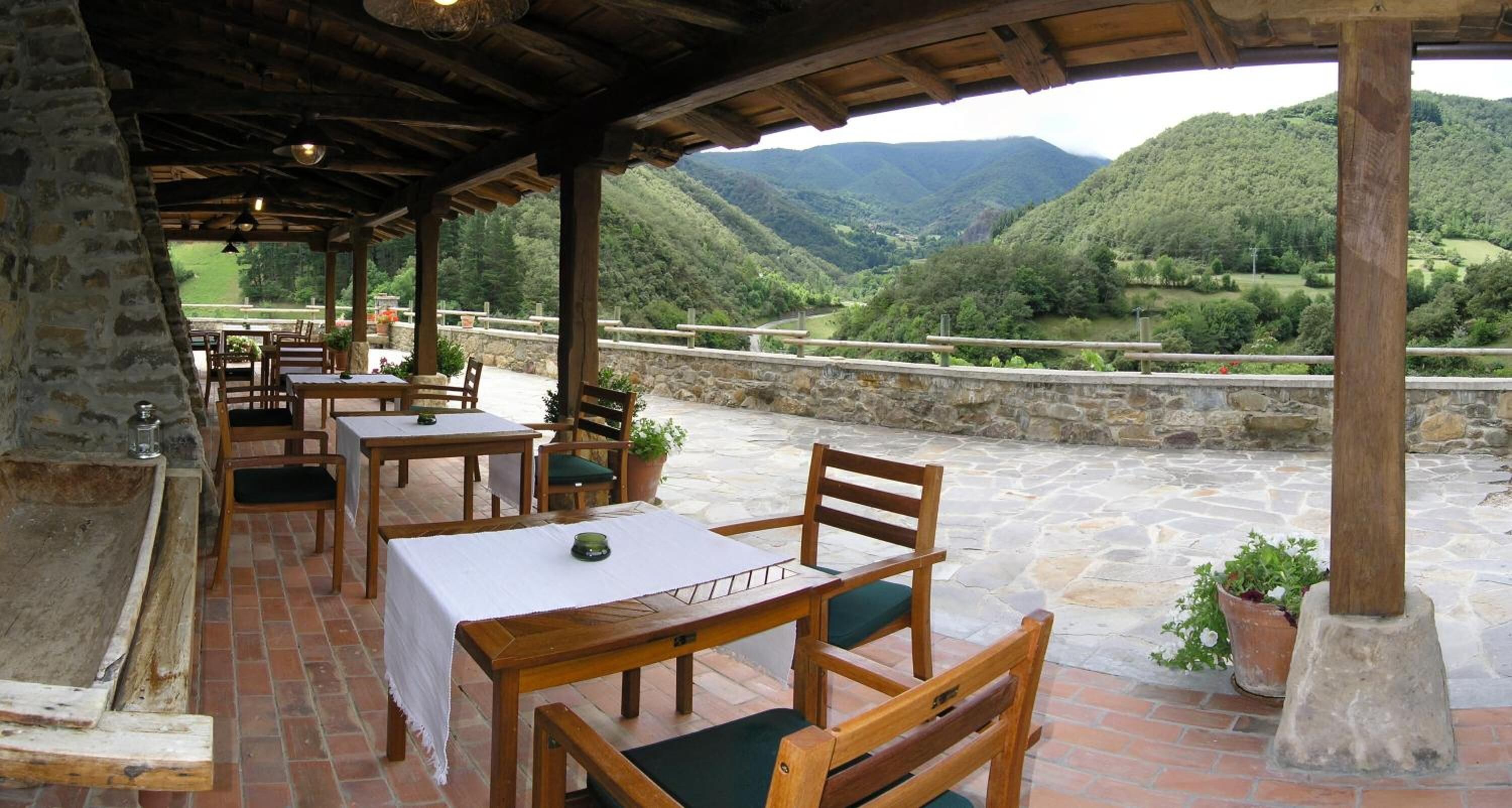 An outdoor patio with stone flooring, wooden tables and chairs, and a view of a mountainous landscape.