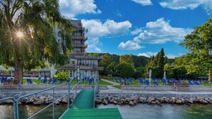 A modern building with a multi-level structure, a green pathway leading to a body of water, and blue lounge chairs arranged in rows. - Világos Hotel Balatonvilágos (Balatonvilagos)