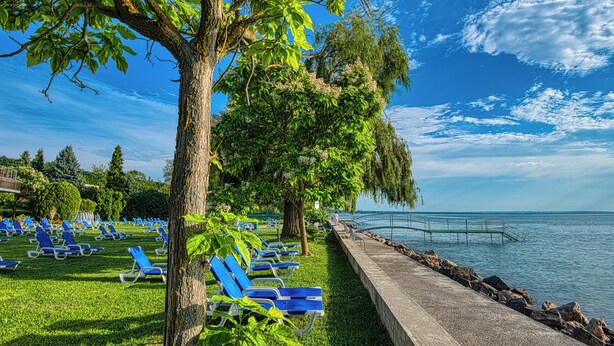 A lakeside park with blue lounge chairs, a concrete walkway, and a stone railing. - Világos Hotel Balatonvilágos (Balatonvilagos)