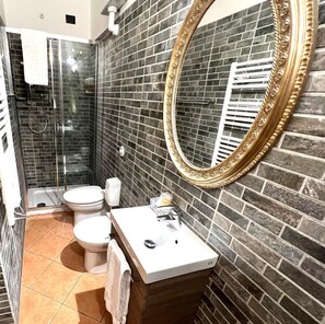 A bathroom with a glass shower enclosure, a white toilet, a sink with a mirror, and a tiled wall. - Virginia's Rooms (Genoa)