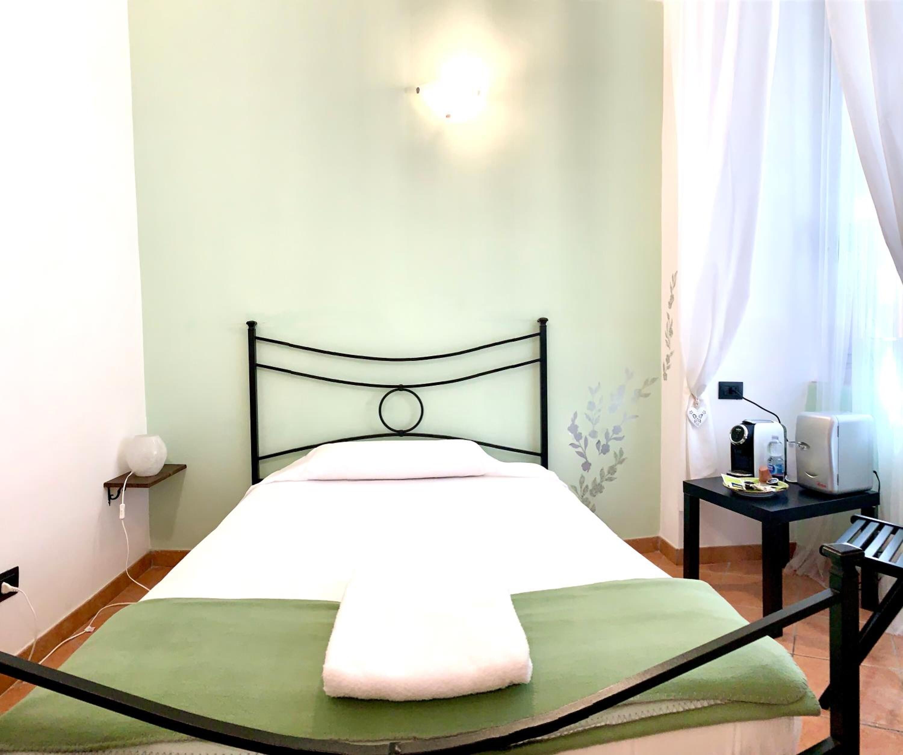A bed with a green bedspread and a white pillow, a black metal headboard, a bedside table with a lamp, and a window with curtains.