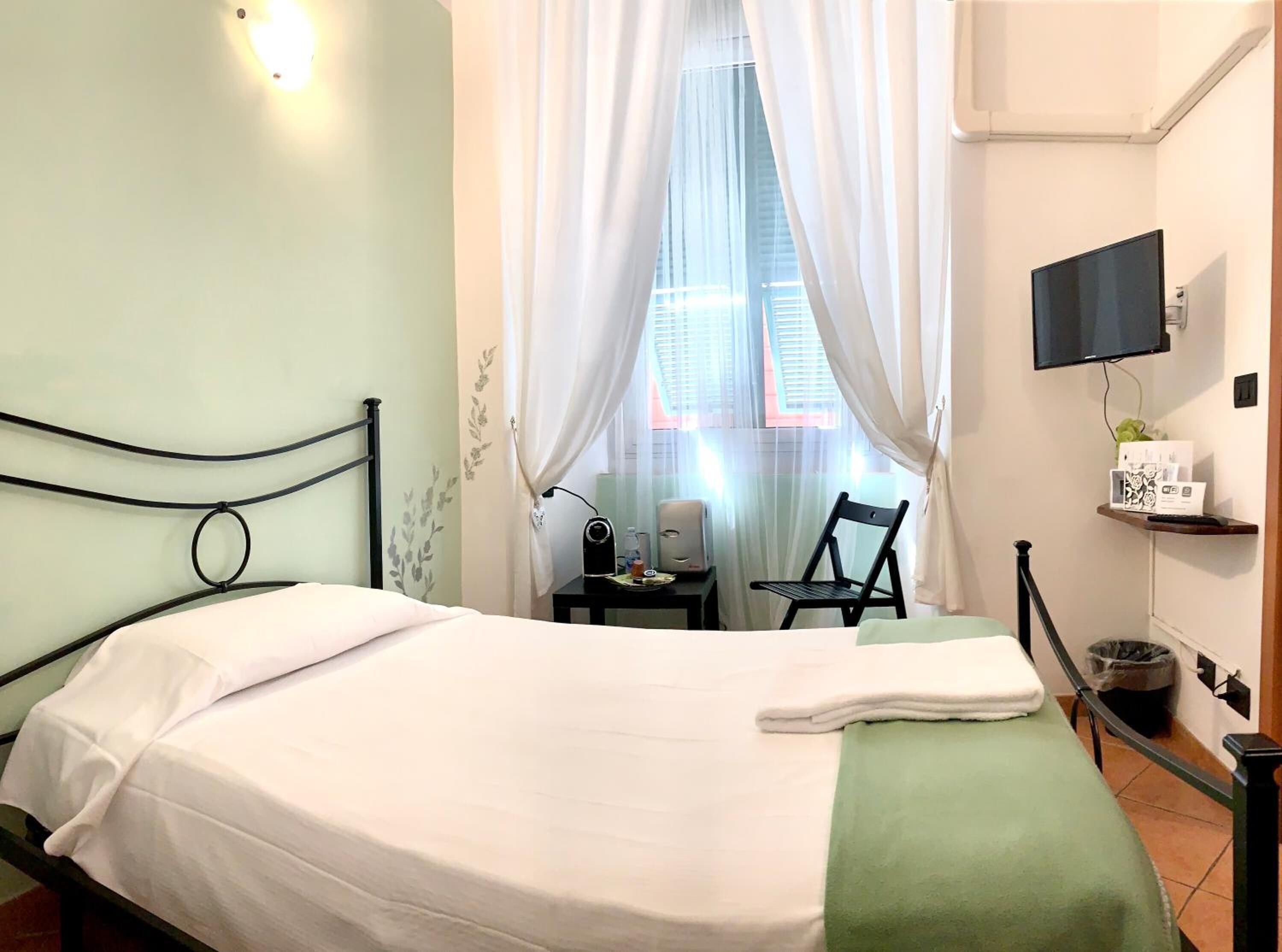 Una camera d'albergo con un letto, una scrivania con una sedia, una TV a parete e una finestra con tende.