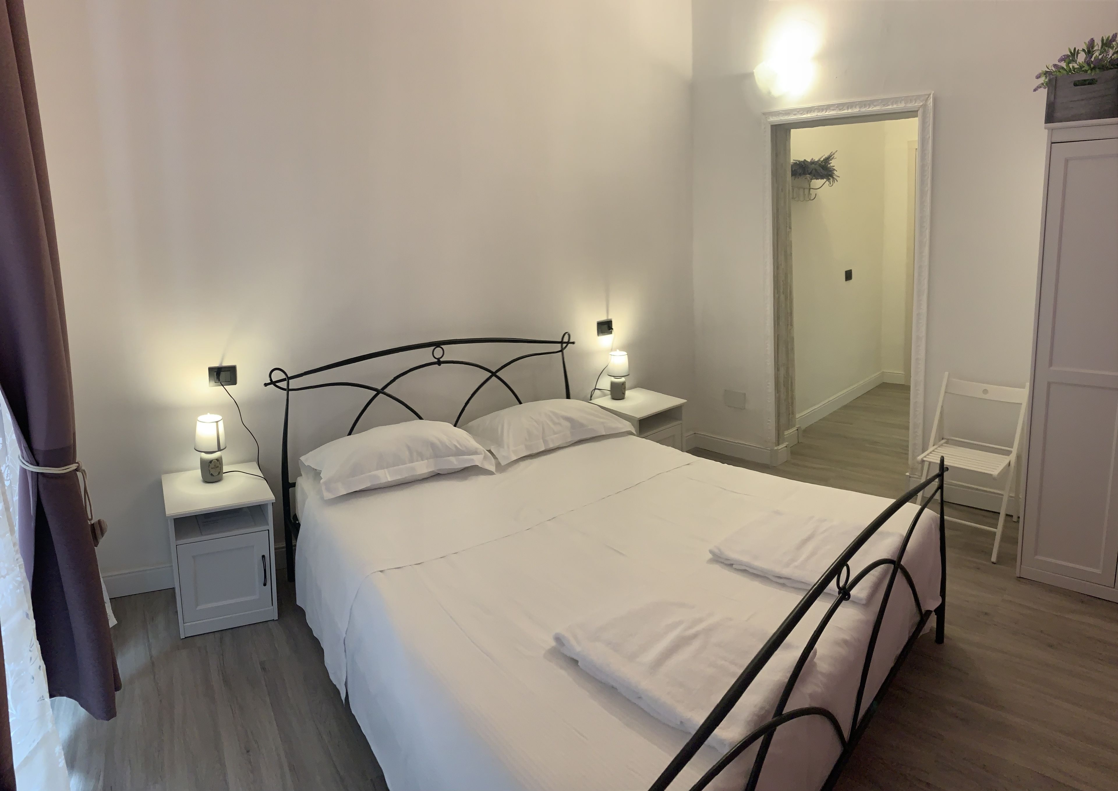 Un lit bien fait, avec du linge de lit blanc et des oreillers, deux tables de chevet avec des lampes, une armoire blanche et un miroir au mur.