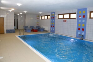 Una piscina interior con paredes de azulejos, agua azul y un área de descanso con cojines azules y rojos.