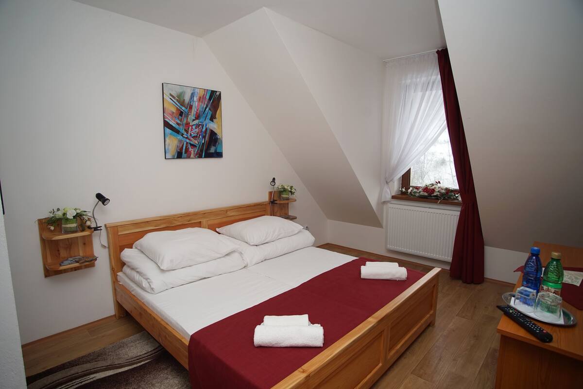 A bedroom with a wooden bed, white linens, a red bedspread, a painting on the wall, a window with curtains, and a bedside table with a remote control and bottles.