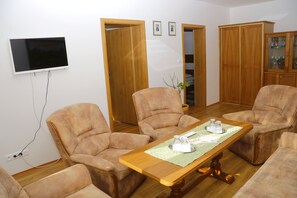 Una sala de estar con una mesa de madera, cuatro sillones color beige, un televisor de pantalla plana montado en la pared y armarios de madera.