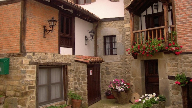 A traditional stone building with a courtyard, featuring a wooden balcony with flower boxes, a stone door, and a small garden area with potted plants. - Casa Reda - Posada de Viñón (Cillorigo de Liebana)