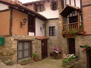 Un bâtiment traditionnel en pierre doté d’une cour intérieure, avec un balcon en bois orné de jardinières fleuries, une porte en pierre et un petit jardin agrémenté de plantes en pot.