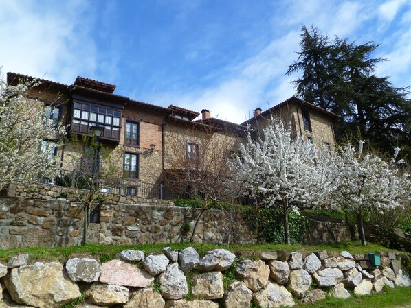 A stone building with multiple windows and balconies, surrounded by a stone wall and flowering trees. - Casa Reda - Posada de Viñón (Cillorigo de Liebana)
