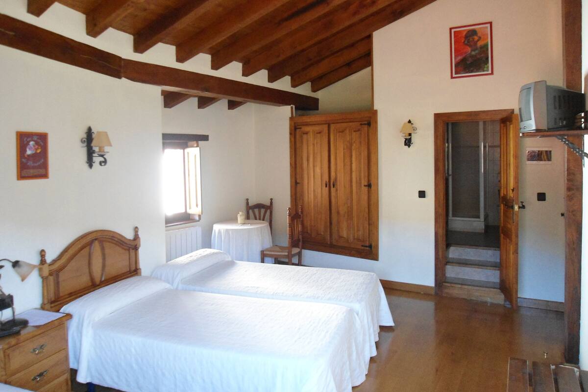 A bedroom with two beds, wooden ceiling beams, a small table with a chair, a wardrobe, and a door leading to another room.