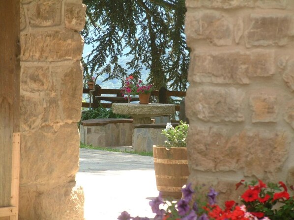 Une arche en pierre, située à l’extérieur, mène à un espace agrémenté d’un banc en pierre, de plantes en pot et de fleurs.