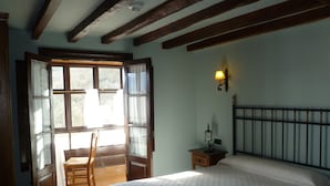 A bedroom with a bed, a chair, a nightstand, a lamp, and a window with curtains. - Casa Reda - Posada de Viñón (Cillorigo de Liebana)