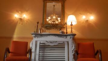 A room with a white ornate fireplace, two red armchairs, a chandelier, and wall sconces.