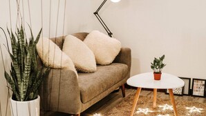 A cozy living room with a brown sofa, fluffy pillows, a white round table, a potted plant, and a floor lamp. - Michi (Hanoi)