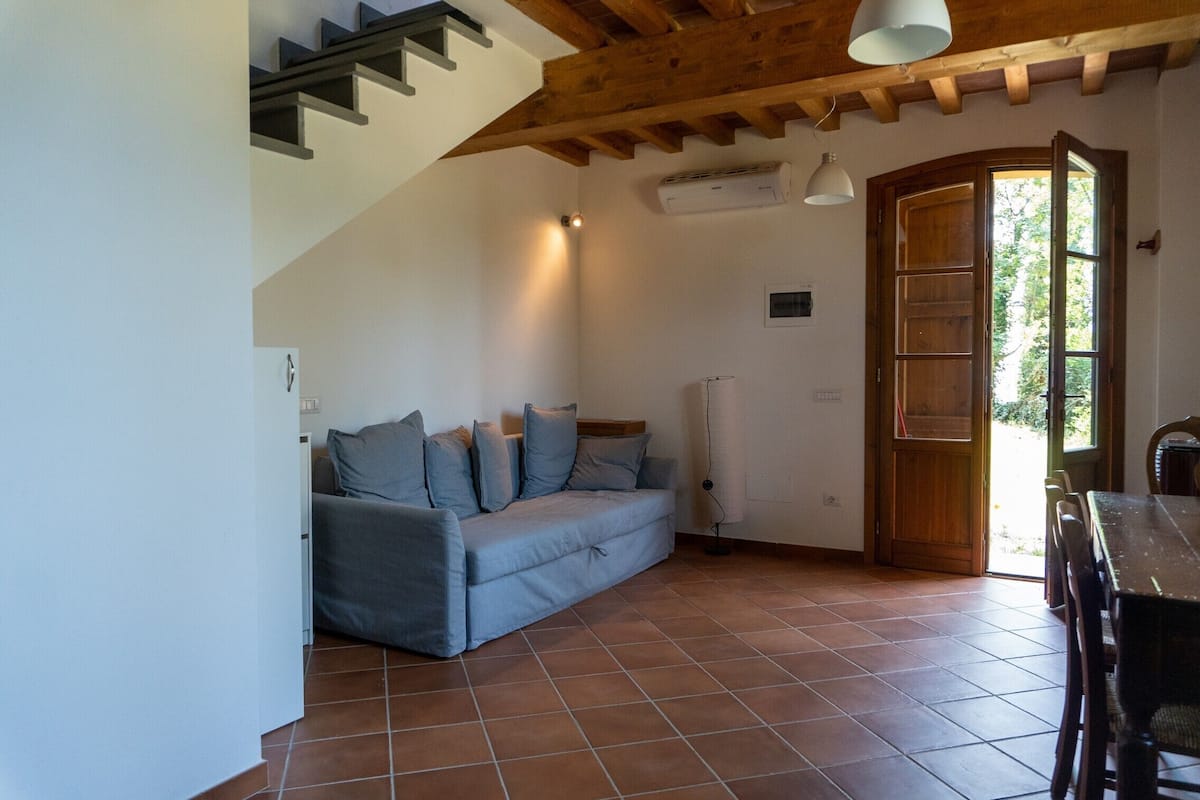 Una habitación con techo de madera, un sofá gris con cojines, piso de baldosas, una puerta de madera y una mesa con sillas.