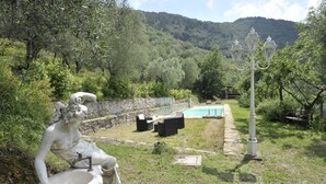 Una estatua de una figura sentada con una pila, mesas y sillas al aire libre, una piscina y un farol en un jardín.