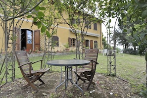 A rustic outdoor seating area with a round metal table and wooden chairs, surrounded by a garden and a yellow building with wooden shutters. - Agriturismo La Grotta (San Giuliano Terme)