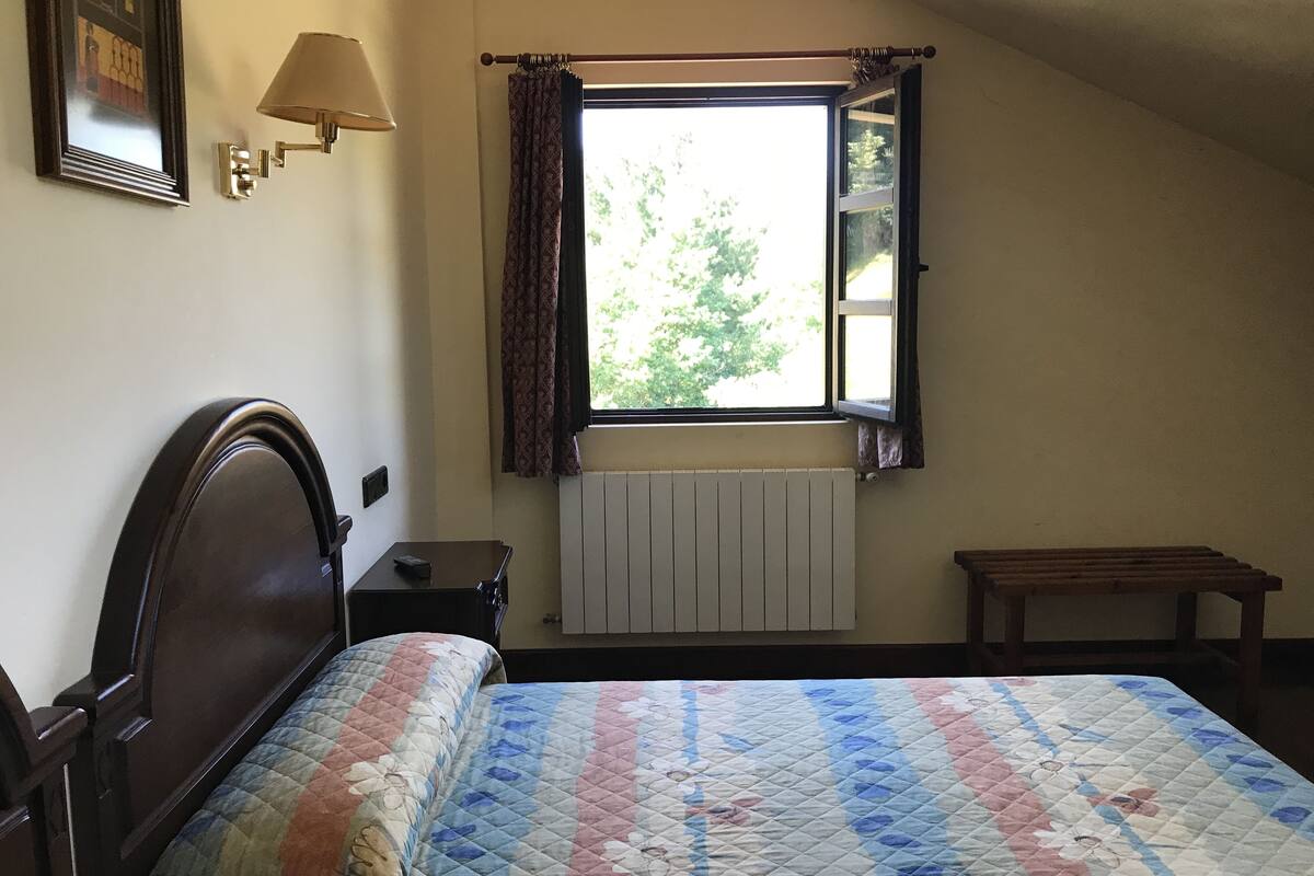 Ein Schlafzimmer mit einem Bett, einem Nachttisch, einem Fenster mit Vorhängen, einem Heizkörper und einem kleinen Holzschemel.