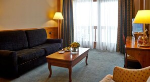A hotel room with a dark-colored sofa, a wooden coffee table with a vase of flowers, a wooden side table with a lamp, and a view through sheer curtains. - Sport Village (Soldeu)