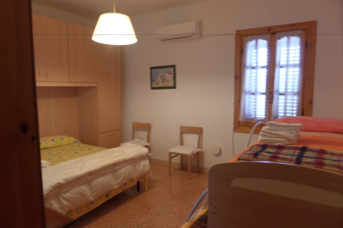 Un dormitorio con dos camas, una silla, una ventana con persianas y una lámpara colgante.