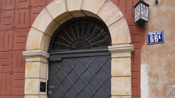 Uma porta de metal preta com padrão de losangos, emoldurada por um arco de pedra.