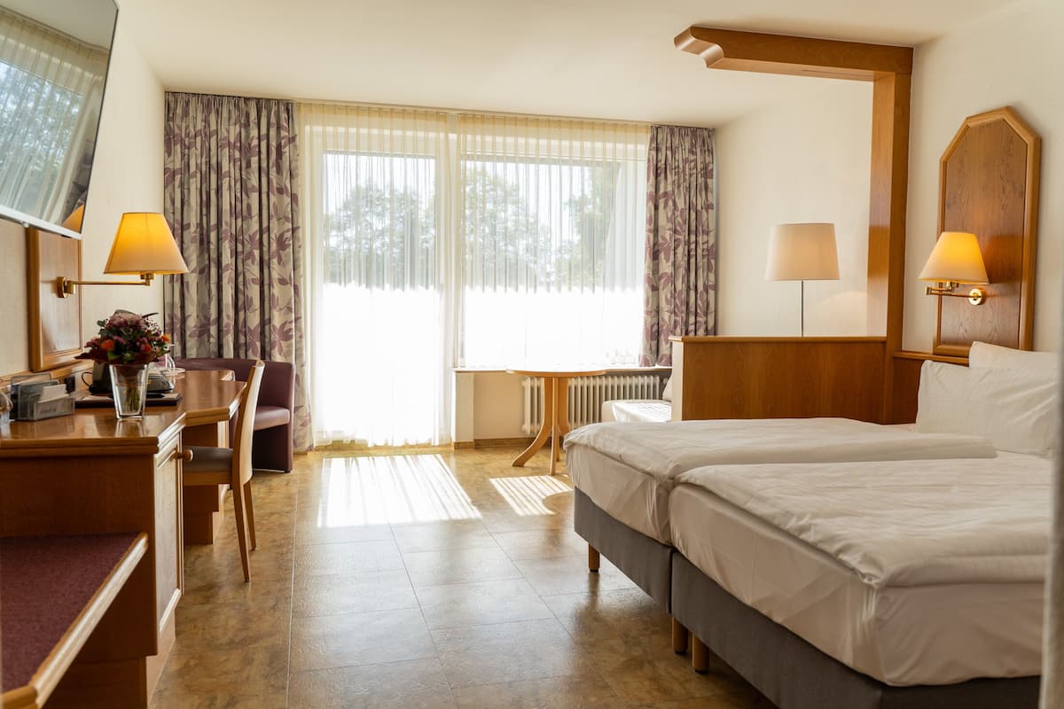 Ein Hotelzimmer mit zwei Betten, einem Schreibtisch mit Lampe, einem Sessel und Blick ins Freie durch große Fenster.