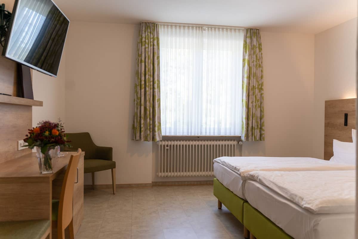 Ein Hotelzimmer mit Bett, Schreibtisch mit Stuhl, Wandfernseher und einem Fenster mit leichtem Vorhang.