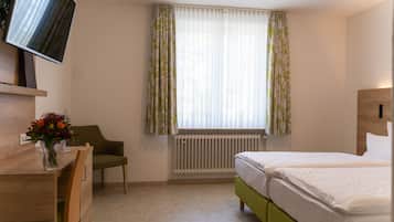 Ein Hotelzimmer mit Bett, Schreibtisch mit Stuhl, Wandfernseher und einem Fenster mit leichtem Vorhang.