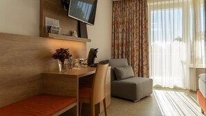 Ett modernt hotellrum med ett skrivbord i trä, en bekväm soffa, en platt-TV som är monterad på väggen och ett stort fönster med tunna, genomskinliga gardiner.