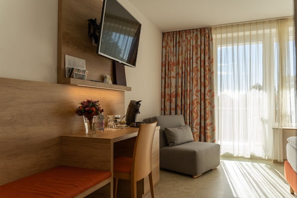 Una habitación de hotel moderna con un escritorio de madera, un sofá cómodo, un televisor de pantalla plana montado en la pared y un ventanal con cortinas traslúcidas.