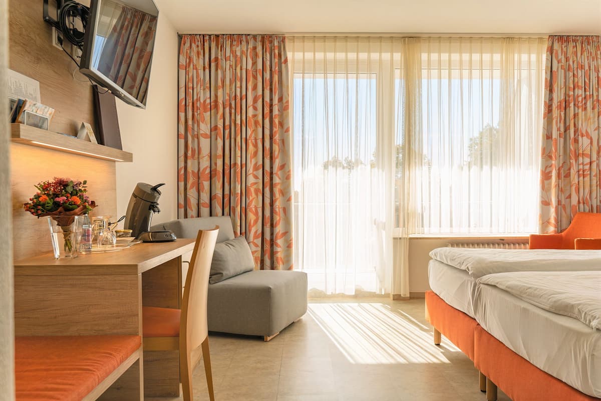 Ein modernes Hotelzimmer mit einem Holztisch, einem bequemen Sessel, einem Bett mit orangefarbener Bettwäsche und einem großen Fenster mit Vorhängen.