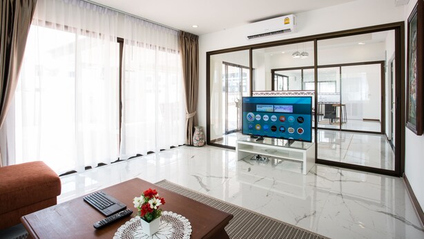 Una habitación de hotel moderna con un televisor de pantalla plana grande, un área de estar confortable con un sofá y una mesa de centro de madera, y un ventanal con cortinas translúcidas.