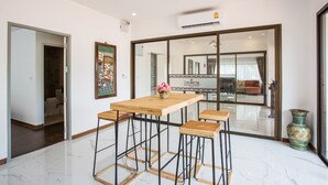 Habitación de hotel moderna con una mesa de madera y taburetes tipo bar, un espejo grande y vistas del área de estar a través de una puerta de vidrio.