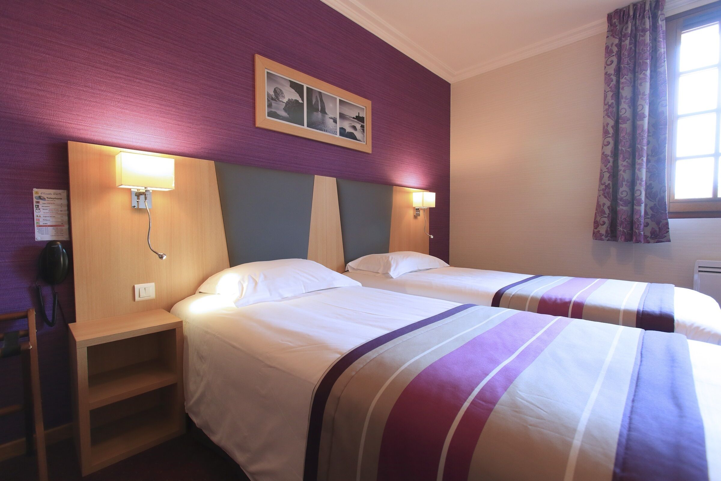A hotel room with two beds, a wooden nightstand, a telephone, and a wall-mounted lamp.