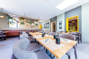 A modern restaurant with long wooden tables, grey upholstered chairs, and hanging plants. - Hôtel L'Escale (Escalles)