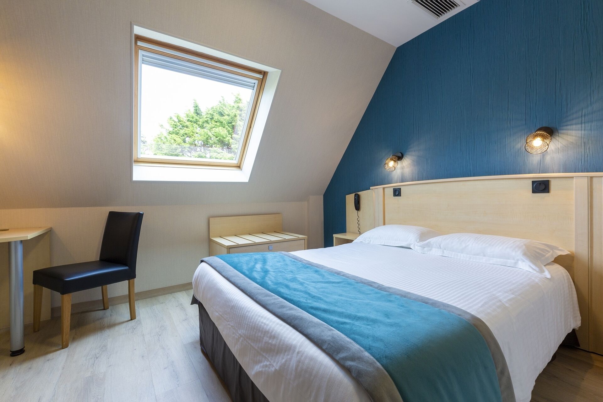 Ein modernes Hotelzimmer mit einem Bett, einem Schreibtisch und einem Stuhl. Ein Fenster lässt natürliches Licht herein und bietet Blick auf Grünfläche.