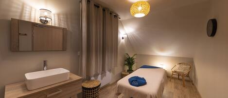 A bedroom with a wooden floor, a bed with a blue towel, a bedside table, a small stool, a potted plant, a wall-mounted clock, and a ceiling light fixture.