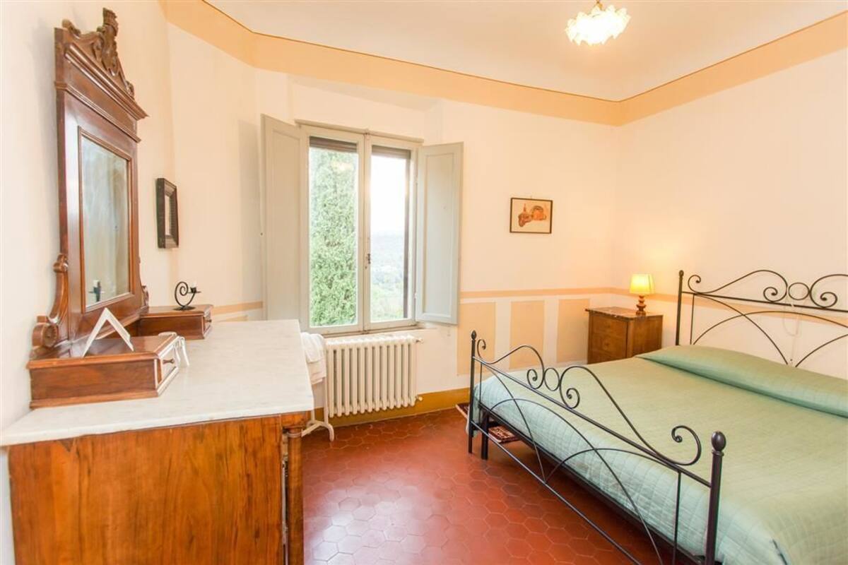Quarto com escrivaninha de madeira, cama com estrutura metálica, janela com vista para vegetação, radiador e mesa de cabeceira pequena com abajur.