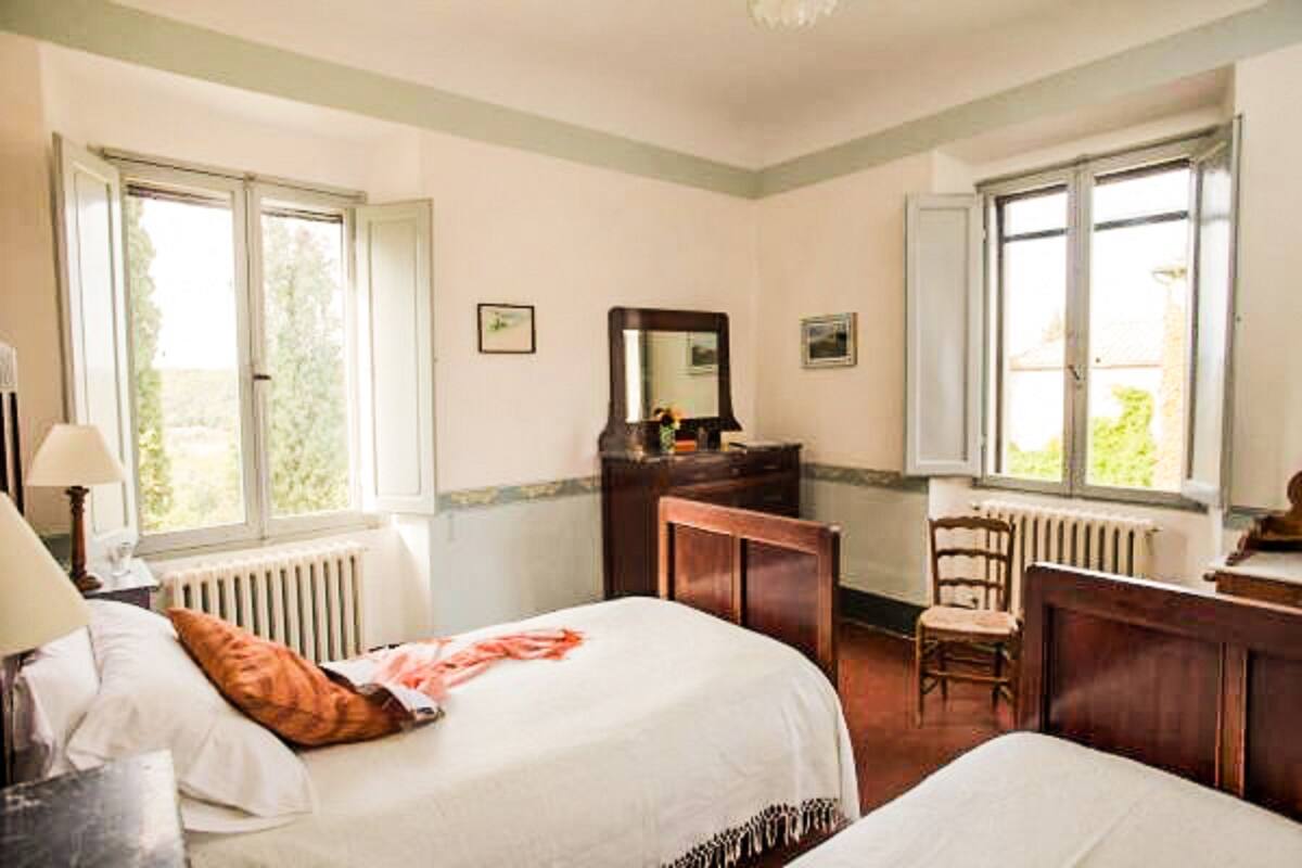 Quarto com duas camas, cômoda, cadeira e janela com vista para o verde.
