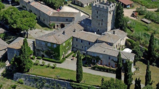 Vista aérea de una finca histórica con varios edificios, incluyendo una torre destacada, rodeada de jardines y árboles bien cuidados.