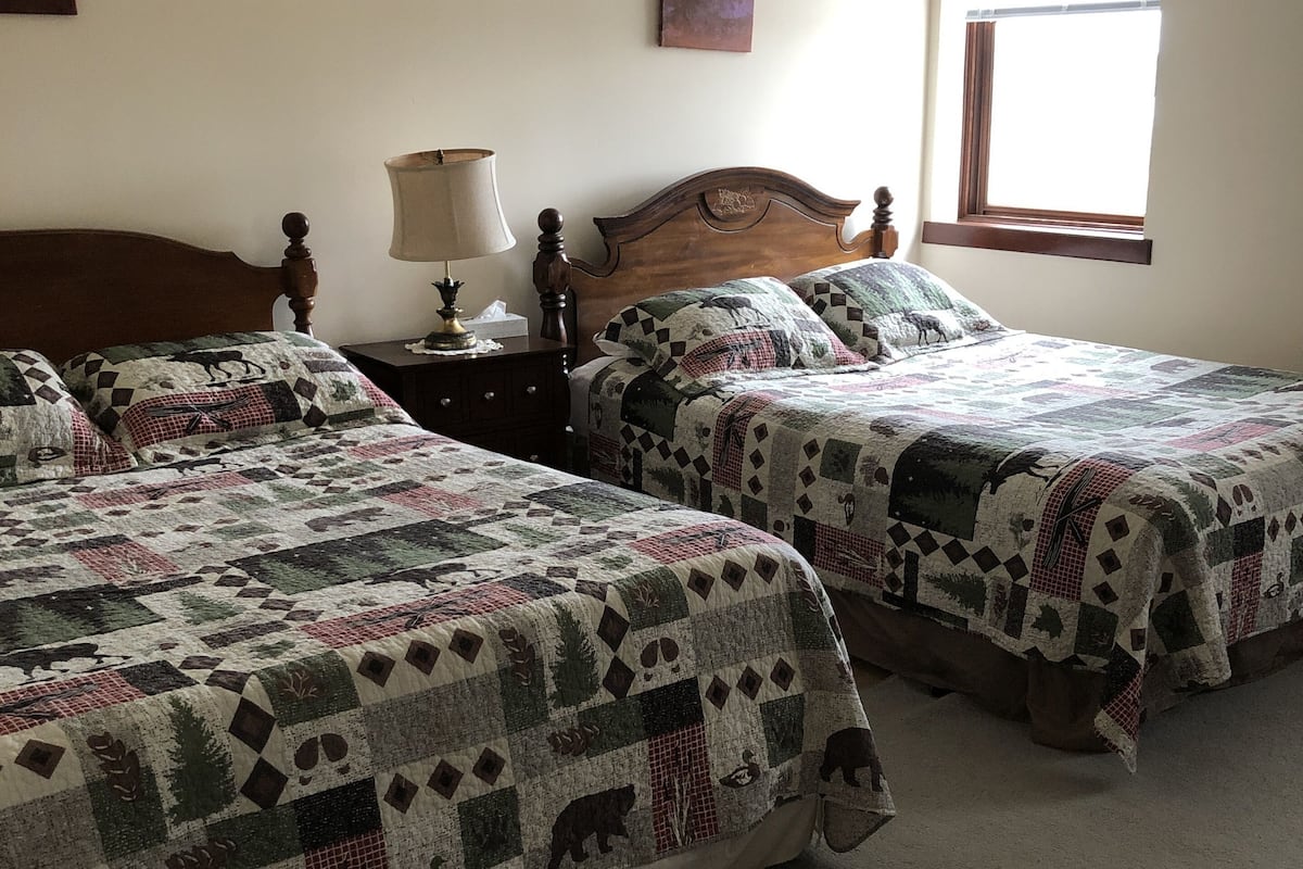 A bedroom with two beds, a nightstand with a lamp, and a window with blinds.