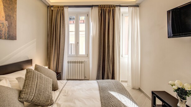 A bedroom with a bed, curtains, a radiator, a TV, and a vase with flowers. - Residenza Costaguti (Rome)