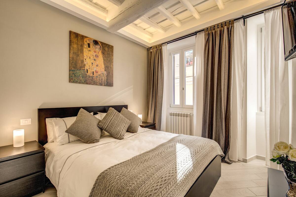 Ein Schlafzimmer mit einem großen Bett, einem Nachttisch, einem Gemälde und einem Fenster mit Vorhängen.