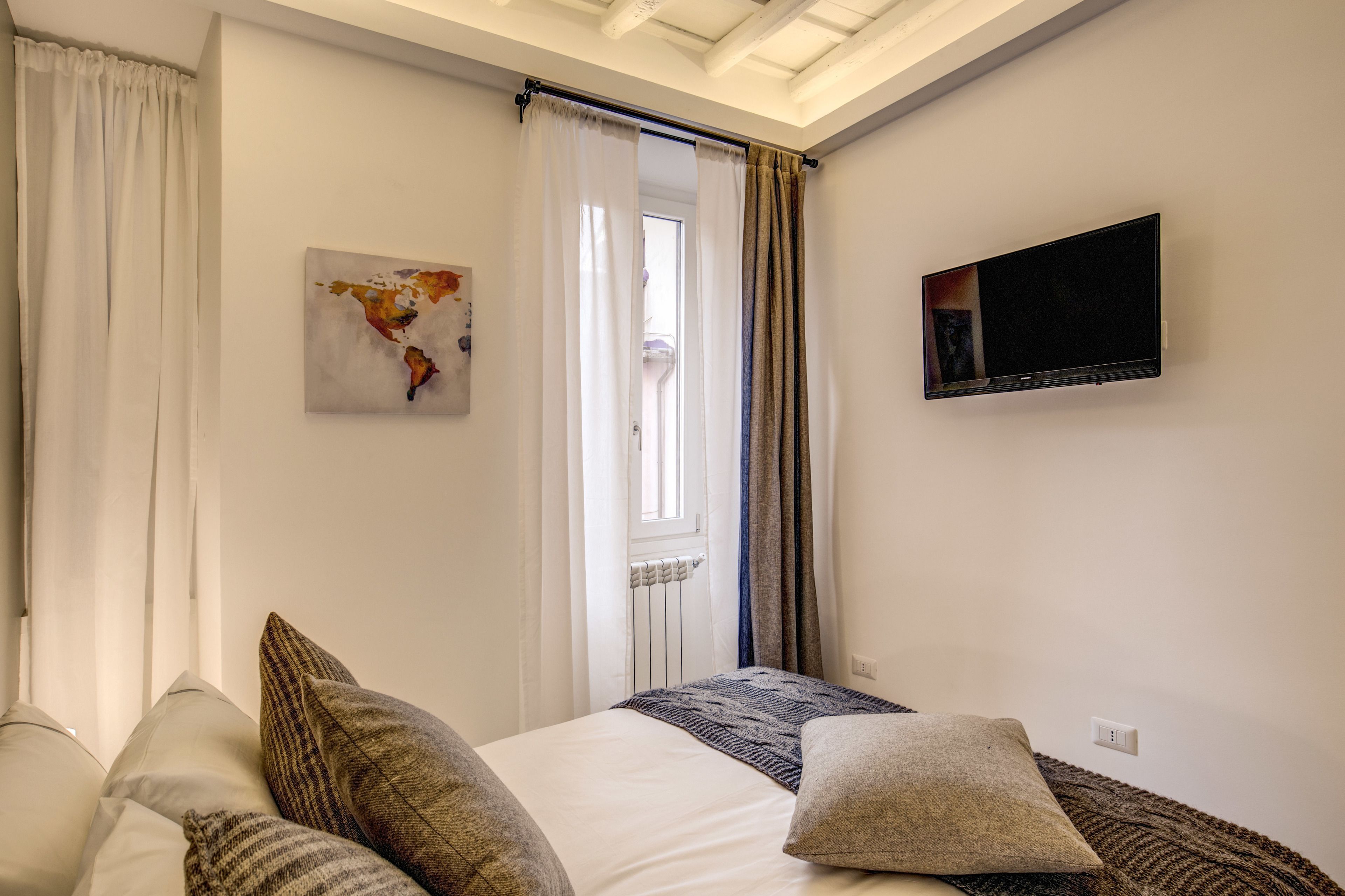 A bedroom with a bed, pillows, a television, a window with curtains, and a wall-mounted artwork.