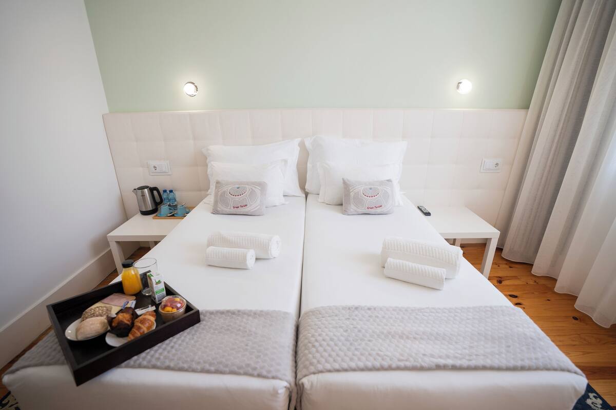 A hotel room with two beds, a small table with a tray of breakfast, and a bedside table with a kettle and bottles.