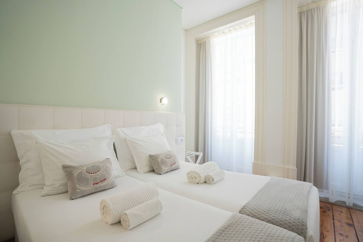 A hotel room with two beds, white bedding, a headboard, and a window with curtains.