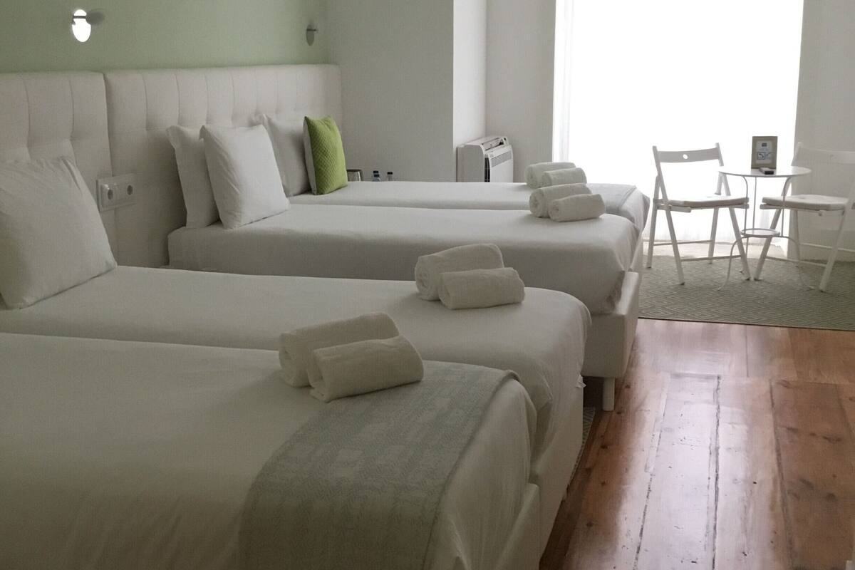 A hotel room with two beds, a small table with chairs, a window with curtains, and a wall-mounted air conditioner.