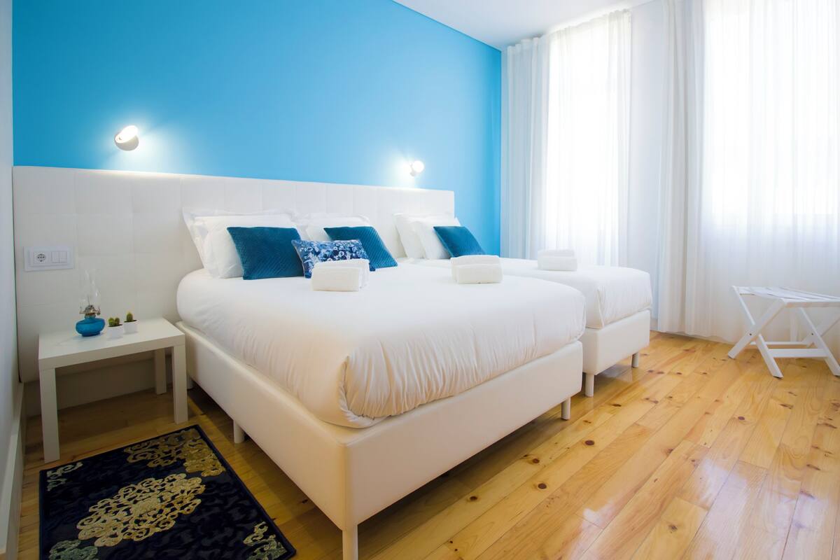 A modern bedroom with a large bed, bedside tables, a small table, and a window with curtains.